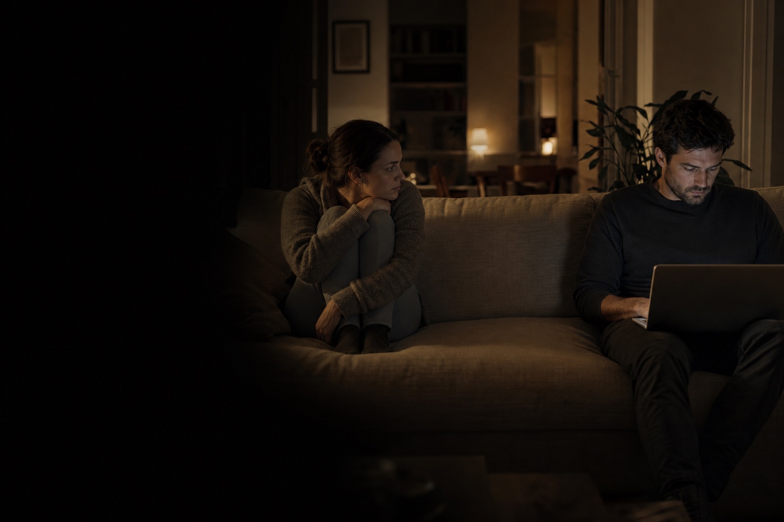 A woman on a sofa watches her partner, who is absorbed in his laptop