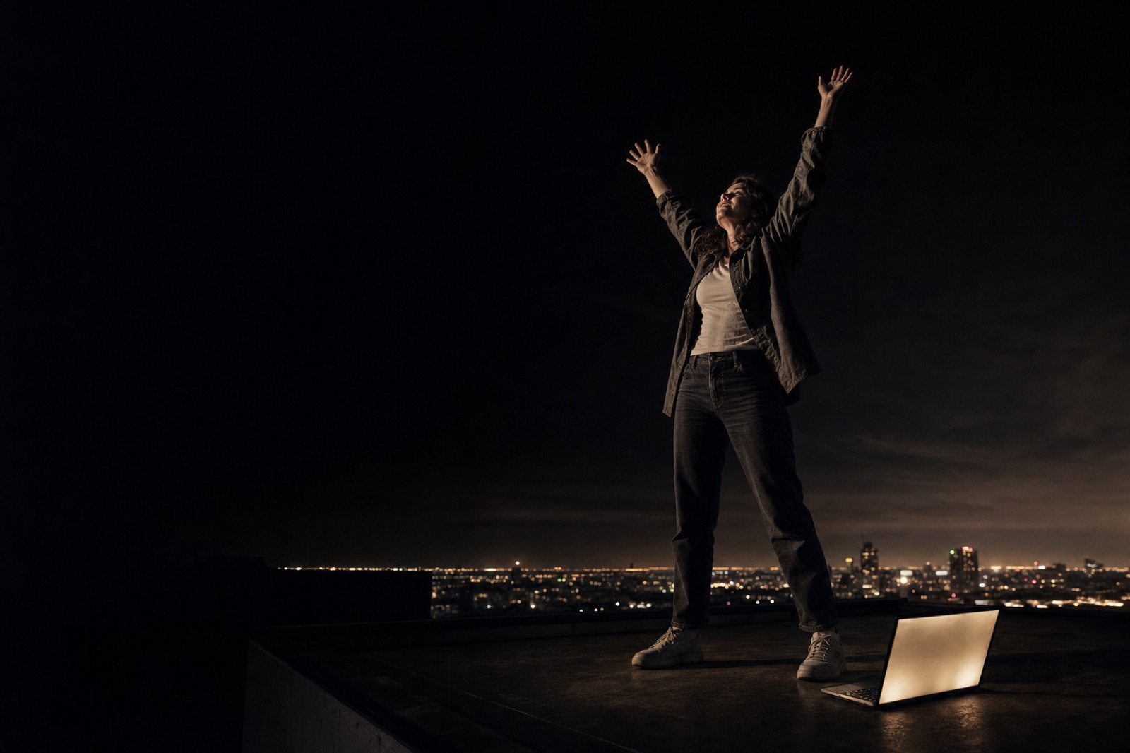 A woman on a rooftop at night with arms raised in triumph, a laptop glowing at her feet