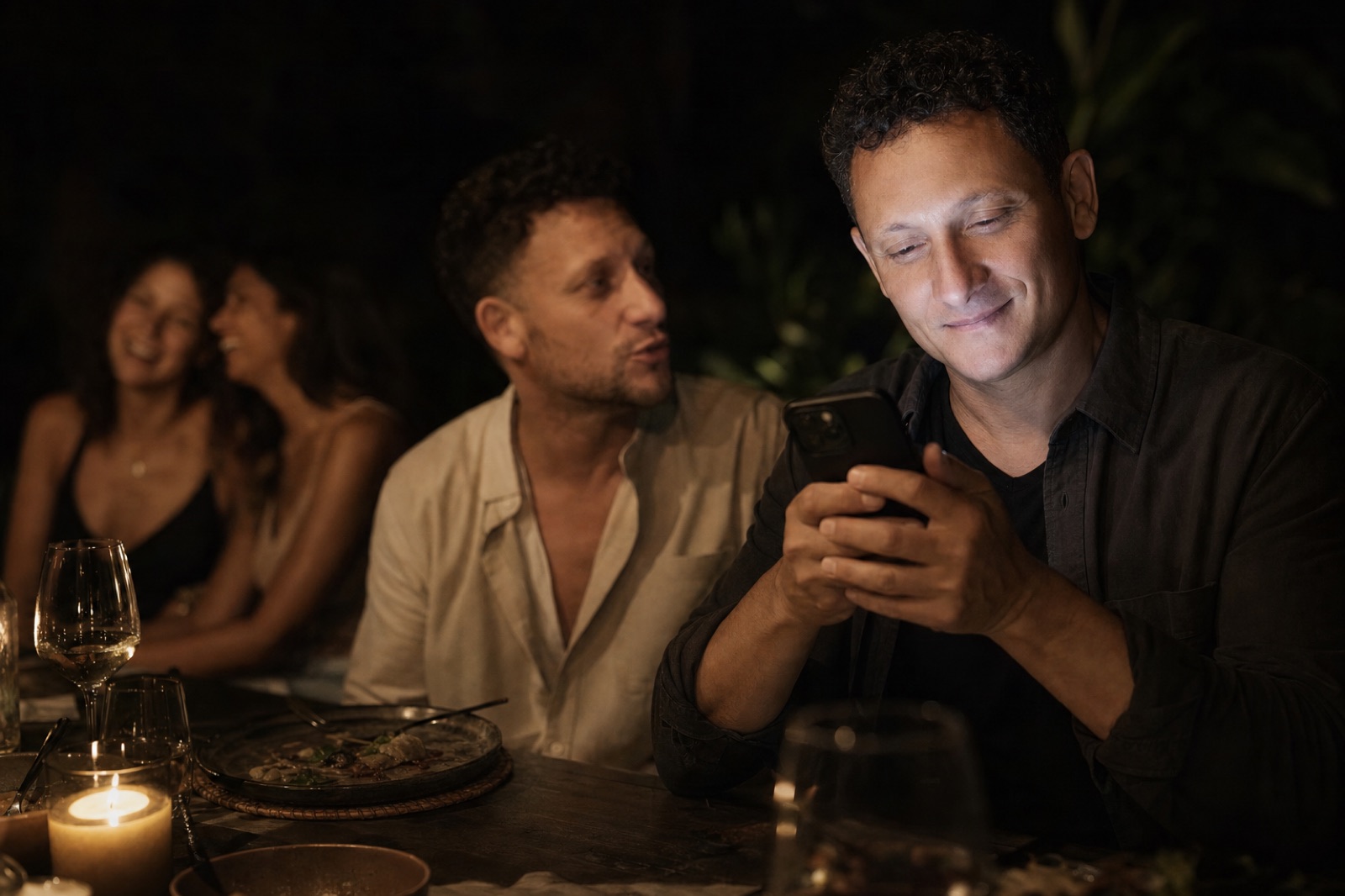 Joe looks down at his glowing phone at a candlelit dinner while a friend beside him talks to him